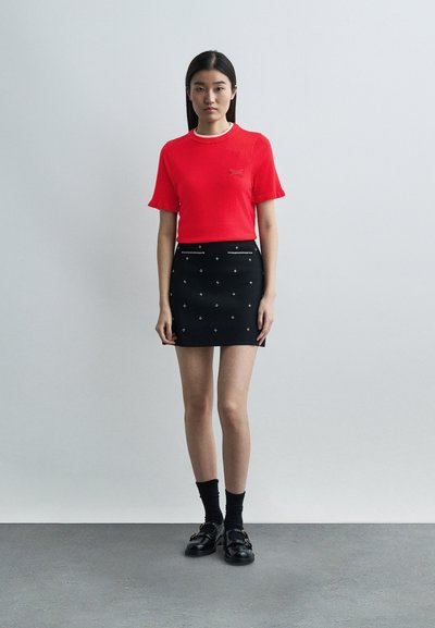 Νεαρή γυναίκα στέκεται δίπλα σε έναν απλό τοίχο, φορώντας κόκκινη μπλούζα με κοντά μανίκια, μαύρη μίνι φούστα με διακοσμητικά, μαύρες κάλτσες και μαύρα παπούτσια.