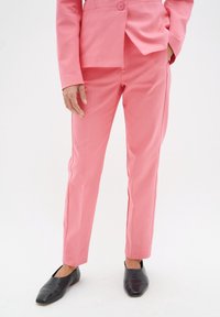 Růžový oblekový sako a odpovídající slim-fit kalhoty, vyrobené z hladké látky. Kalhoty mají čistý střih, bez viditelných vzorů nebo ozdob.