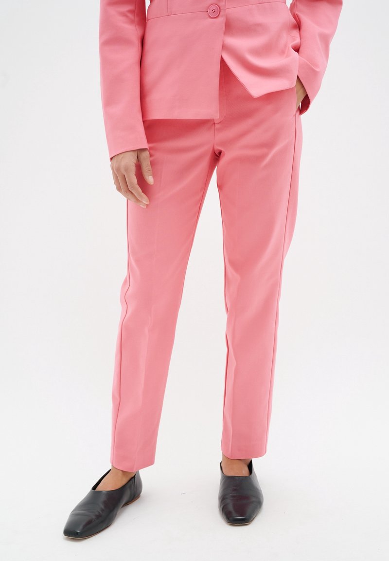 Růžový oblekový sako a odpovídající slim-fit kalhoty, vyrobené z hladké látky. Kalhoty mají čistý střih, bez viditelných vzorů nebo ozdob.