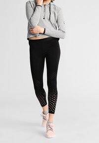 Osoba v seříznuté šedé mikině, černých legínách s perforovanými vzory na lýtkách a růžových sportovních botách, stojí na jedné noze.