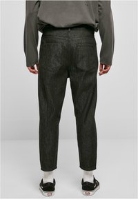 Pantalon en denim gris foncé avec une coupe droite, doté de poches arrière et d'une texture subtile. Porté avec des chaussures noires et des chaussettes blanches.