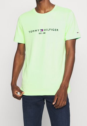 Man draagt een neon groene Tommy Hilfiger T-shirt met logo en de tekst "EST. 1985", gecombineerd met donkerblauwe jeans tegen een effen achtergrond.