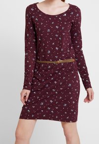 Langärmliges, kastanienbraunes Kleid mit kleinen Blumenmustern. Verfügt über einen runden Halsausschnitt und einen geflochtenen braunen Gürtel in der Taille.