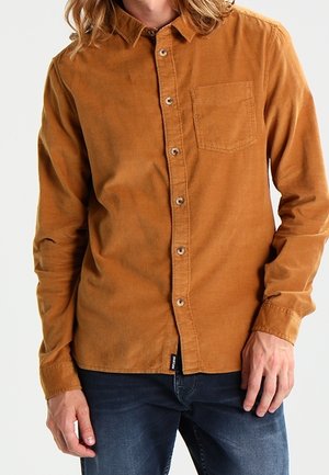 Homme portant une chemise en velours côtelé orange brûlé à manches longues, boutonnée, avec une poche poitrine et un jean bleu foncé, debout devant un fond uni.