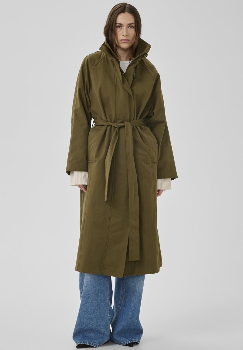 Vrouw draagt een olijfgroene trenchcoat met riem over een witte trui en wijde blauwe jeans, staand tegen een effen lichte achtergrond.