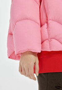 Roze pufferjack met een textuur, gequilt ontwerp. Heeft een rode geribde tailleband. Een deel van een hand is zichtbaar bij de manchette, rustend tegen donkere stof.