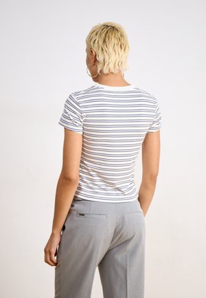 Persoon met kort blond haar, gouden oorringen, een wit shirt met korte mouwen met dunne zwarte strepen en lichtgrijze broek, staande met de rug naar de camera.