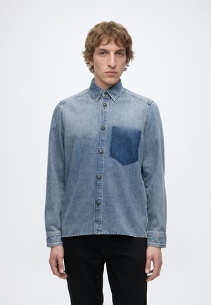 Jeune homme aux cheveux bouclés portant une chemise en denim bleu délavé avec une poche poitrine bleu plus foncé et un pantalon noir, debout devant un fond uni.