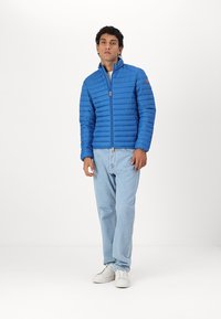 Blauwe pufferjack met horizontale quiltpatroon, volledige ritssluiting en staande kraag. Draagt het met lichtblauwe jeans en witte sneakers.