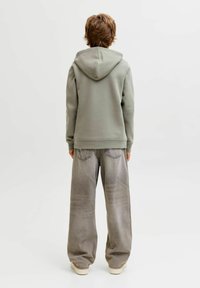 Svetlo zelena majica s kapu, v kombinaciji z ohlapnimi svetlo sivimi kavbojkami. Mehka tekstura, sproščen kroj, z vidnimi zadnjimi žepi na kavbojkah.