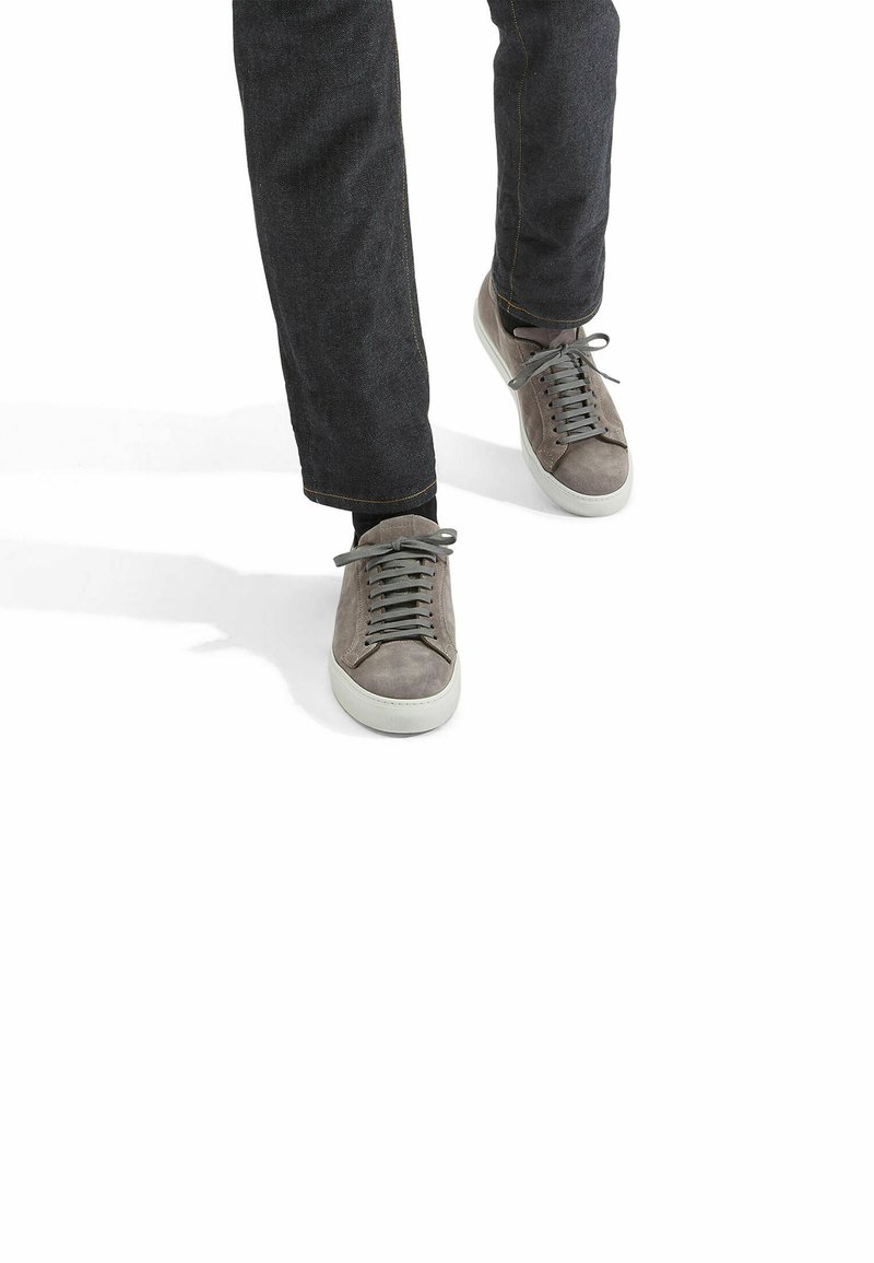 Graue Wildledersneaker mit einer weißen Gummisohle, ausgestattet mit grauen Schnürsenkeln und runder Zehenpartie, kombiniert mit einer dunklen Jeanshose mit gelben Nähten.