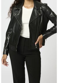 Kvinna iförd en svart läderjacka med silverblixtlås över en vit ribbad topp och svarta byxor, poserar med en hand på höften.