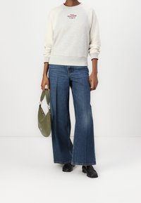 Light grey sweatshirt with raglan sleeves and red embroidered logo, paired with wide-leg blue jeans and a green handbag. Black loafers complete the outfit.