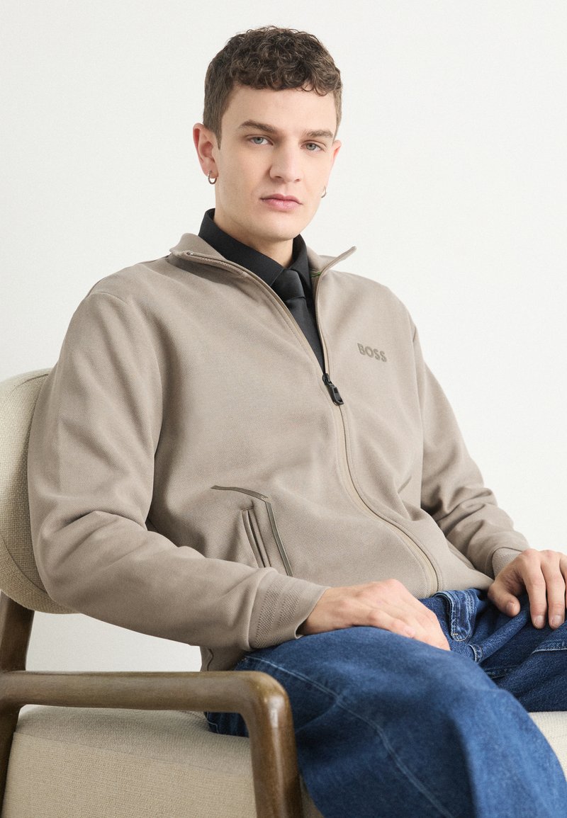 Siva zip-up jakna z teksturiranim zaključkom, ki vsebuje subtilen logotip in kontrastno zadrgo. Nošena preko črne majice in modrih kavbojk.