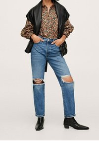 Jeans z wysokim stanem w kolorze niebieskim, z dziurami na kolanach, wystylizowane z koszulą w kwiatowy wzór oraz czarną skórzaną kamizelką. Czarne botki na kostkę, na jednolitym tle.