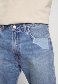 Ljusblå denimjeans med en blekt finish, med en ficka som har en ljusare blå lapp, bältesöglor och metallknäppning.