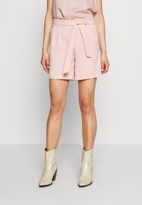 Pantaloni corti su misura rosa chiaro con una cintura, dotati di due tasche laterali, abbinati a stivali alla caviglia beige con texture. Tessuto morbido, design strutturato.