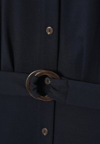 Rochie bleumarin cu un bretele din material, având o cataramă mare, rotundă, de culoare maro închis. Cinci butoane într-o nuanță asortată pe față. Textură netedă.