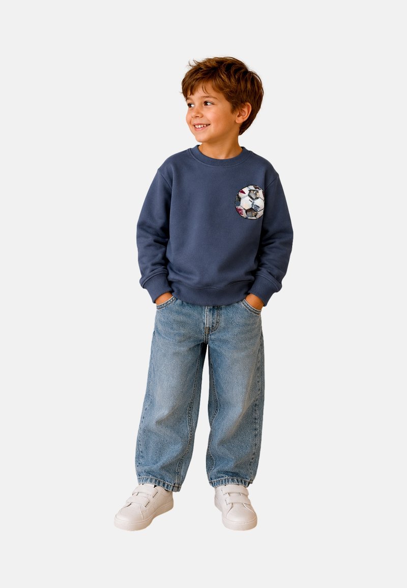 Jeune garçon portant un sweat-shirt bleu avec un motif ballon de football, jeans bleus larges, baskets blanches, mains dans les poches, souriant et regardant vers la droite.