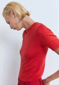 Röd stickad kortärmad tröja med rund halsringning. Slät textur med figursydd design. Modellen står på sidan, med huvudet något sänkt.
