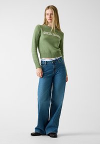 Maglione verde lavorato a maglia con testo "GUESS JEANS", abbinato a jeans larghi blu. Il maglione ha un orlo a costine, i jeans hanno una finitura classica.