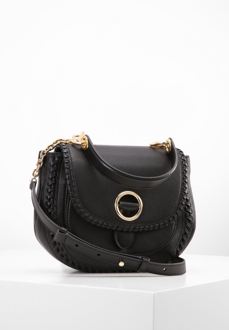Sac bandoulière en cuir noir avec fermoir circulaire doré, bords tressés, poignée courte et bandoulière réglable sur surface blanche.