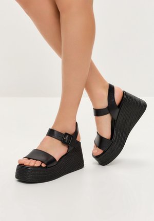 Beine in schwarzen Leder-Keilabsatzsandalen mit breiten Riemen und Knöchelriemen vor weißem Hintergrund, ein Fuß leicht angehoben.