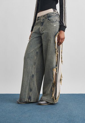 Személy széles szárú szürke farmert visel, fehér rojtos oldalsávokkal és ezüst cipőkkel, kék szőnyegen áll egyszínű háttér előtt.