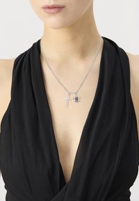 Kvinne som har på seg et sølvkjede med kors- og fleur-de-lis-pendanter over en svart dyp V-hals topp.