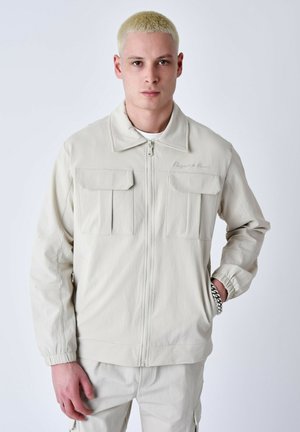 Veste beige clair avec un col, deux poches avant supérieures et des poignets élastiques. Tissu lisse avec une texture subtile et fermeture éclair.