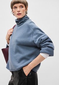 Svetlo modra, udobna turtleneck pulover z širokimi rokavi in rebrastimi manšeti. Ujemajo se z črnimi usnjenimi hlačami, ki jih dopolnjuje bela srajca, vstavljena v hlače.