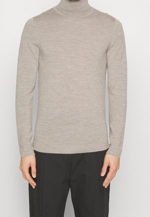 Pull beige à col roulé et manches longues porté avec un pantalon noir sur une personne debout devant un fond clair.