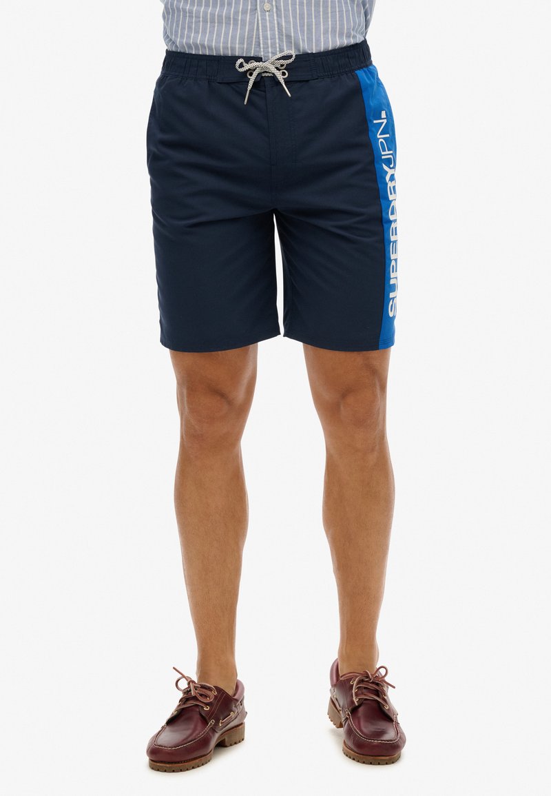 Shorts de bain navy avec un panneau latéral bleu, taille élastique et cordon argenté. Présente un logo imprimé verticalement en blanc.
