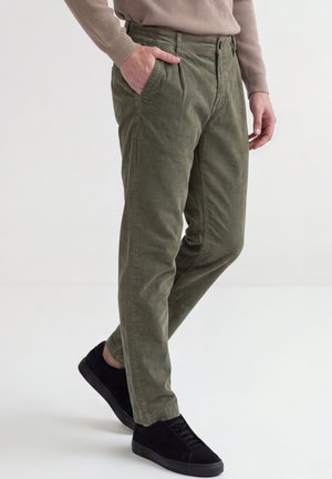 Homme portant un pantalon en velours côtelé vert olive, une main dans la poche, associé à des baskets noires et un pull beige.