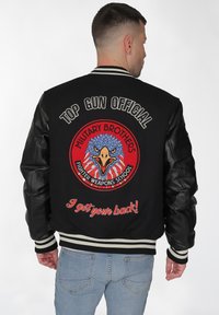 Svart bomberjacka med läderärm, med en stor broderad örn-emblem och texten "TOP GUN OFFICIAL" i vitt och rött.