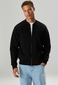 Giubbotto bomber nero con zip frontale, colletto rotondo e polsini a costine. Abbinato a jeans denim azzurri, con una vestibilità rilassata.