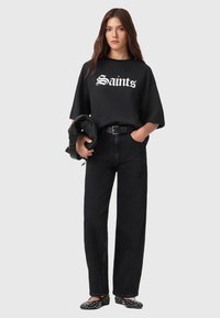 Zwarte oversized t-shirt met de tekst "Saints", zwarte high-waisted jeans en zwarte gespte ballerina's. Model houdt een zwarte jas vast.