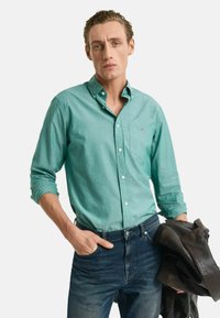 Türkises Button-Down-Shirt mit langen Ärmeln, Taschendetails und Jeans. Das Model hält eine braune Lederjacke.
