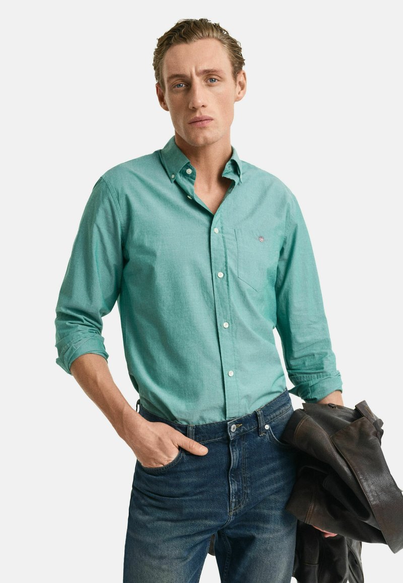 Türkises Button-Down-Shirt mit langen Ärmeln, Taschendetails und Jeans. Das Model hält eine braune Lederjacke.