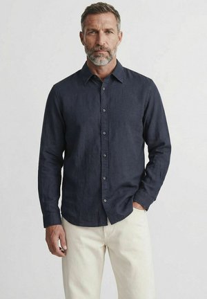 Homme barbu portant une chemise à manches longues bleu marine foncé boutonnée et un pantalon beige clair, debout devant un fond clair uni.