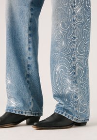 Lichtblauwe denim jeans met decoratieve rhinestone spiralen langs de buitenste naden, gecombineerd met zwarte leren cowboylaarzen.