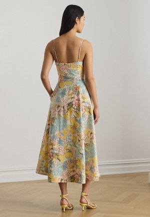 Γυναίκα που στέκεται μέσα σε εσωτερικό χώρο, φοράει ένα αμάνικο midi φόρεμα με floral σχέδια και λεπτές τιράντες, καθώς και κίτρινες σανδάλια με χαμηλό τακούνι, πάνω σε ξύλινο πάτωμα.