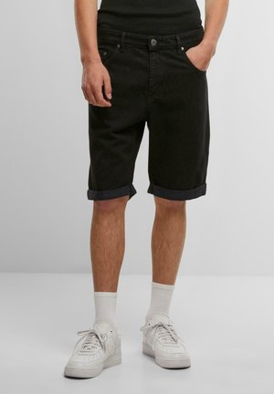 Shorts de denim negro con dobladillos, cinco bolsillos y herrajes metálicos. Combinados con zapatillas blancas y calcetines acanalados visibles.