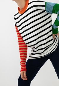 Personne portant un pull rayé avec un devant noir et blanc, une manche gauche rouge et orange, et une manche droite bleue et verte, associé à un pantalon foncé.