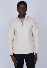 Pull en polaire beige avec un col montant et une demi-fermeture éclair. Coupe ajustée avec des manches longues, associé à un pantalon noir à rayures.