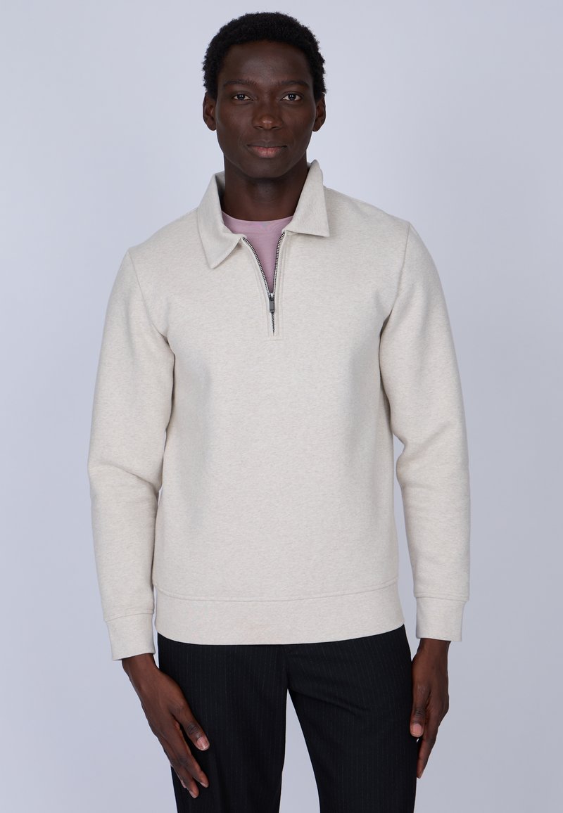 Pull en polaire beige avec un col montant et une demi-fermeture éclair. Coupe ajustée avec des manches longues, associé à un pantalon noir à rayures.