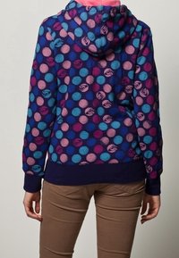 Sweat à capuche avec une base bleu foncé et des pois multicolores en rose, violet et turquoise. Présente des poignets et un ourlet côtelés.