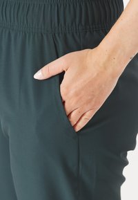 Mörkgröna träningsbyxor med elastisk midja, som har en sidoficka och ett slätt, lätt tyg. Närbild av hand i fickan.