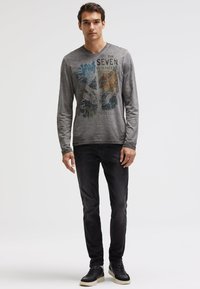 Langärmliges graues T-Shirt mit einem grafischen Druck von Bergen, kombiniert mit dunklen Jeans und schwarzen Sneakers, vor einem schlichten Hintergrund.