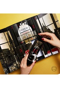 Schwarzer und goldener Adventskalender mit Fenstermotiven, der rote Akzente und Zahlen zeigt. Hand öffnet ein Fach und enthüllt ein kleines Produkt.